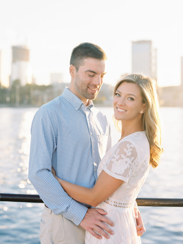 A couple in Charlestown, MA doing their engagement photos along the waterfront. He's in a blue button down shirt and she's in a white dress.
